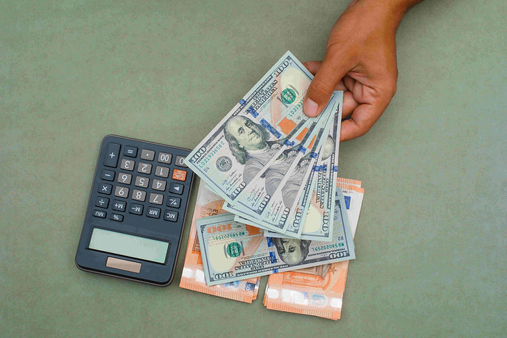 A hand holds multiple $100 bills next to a calculator on a green surface, conveying a financial or budgeting theme.