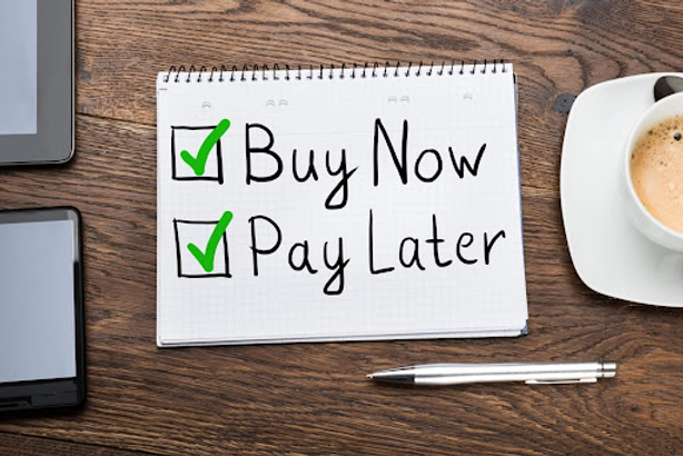 A notepad reading 'Buy Now, Pay Later' with green check marks, on a wooden desk beside a coffee cup and pen.