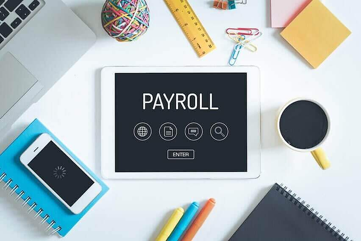 Tablet displaying "PAYROLL" on desk with office items: coffee, notebook, phone, and colorful stationery. Bright, organized workspace.
