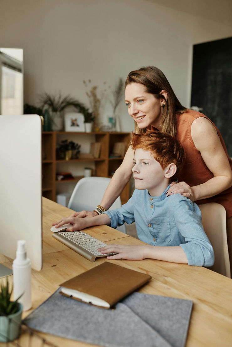 Woman and boy at a desk, using a computer. The woman smiles, hands on boy’s shoulders. Wooden shelves and decor are in the background.