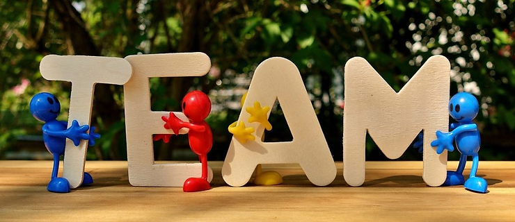 Colorful figures support large white letters spelling TEAM on a wooden table, with greenery in the background, conveying teamwork.