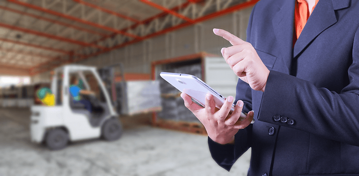 A businessman in a suit using a tablet in a warehouse, with a forklift in the background.