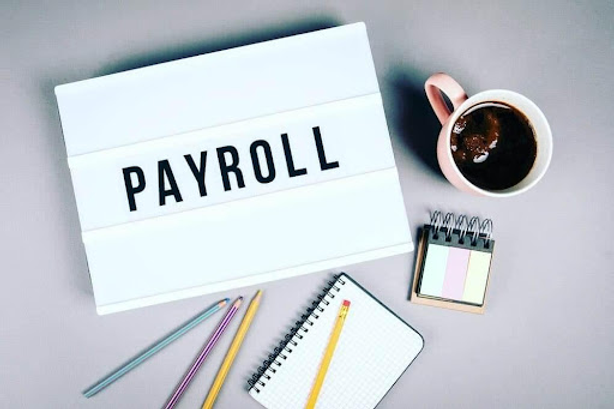 A lightbox sign reading 'Payroll' on a desk with a cup of coffee, colored pencils, sticky notes, and a notebook.