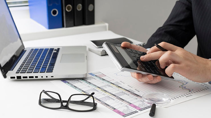 Hands holding a calculator near a laptop, papers with charts, glasses, and a magnifying glass. Office setting with a focused mood.