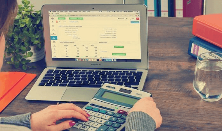 A woman using a calculator next to a laptop showing accounting software at a desk.